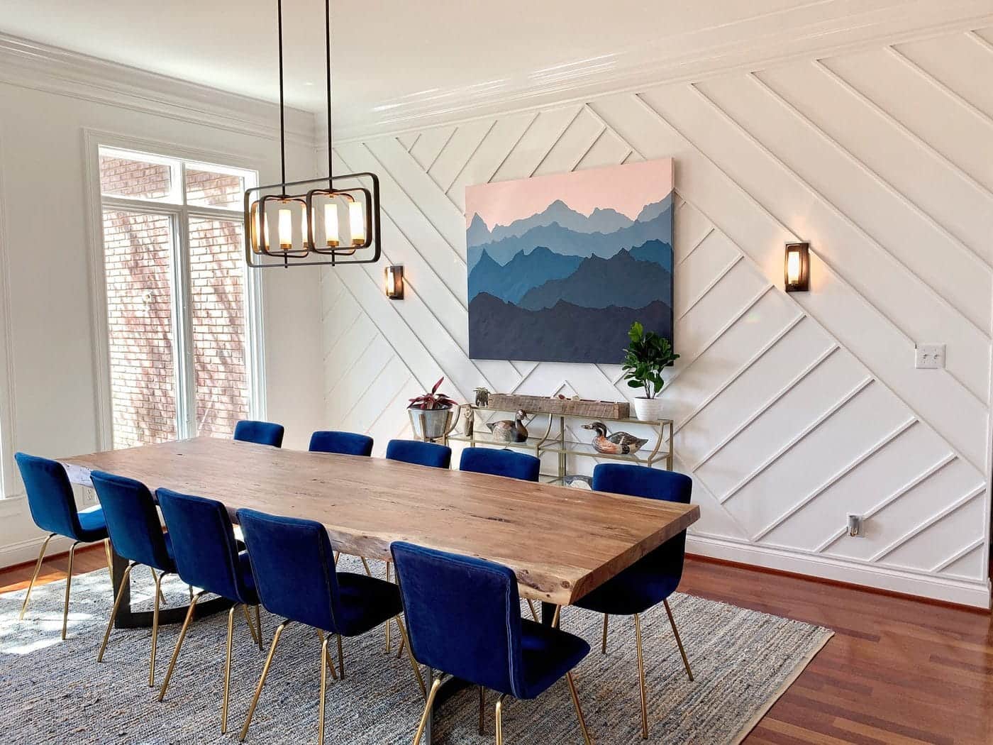 4 Benefits of Implementing a Handmade Dining Table in Your Dining Space. Great panelled accent wall with a painting for large dining room with dark blue plush chairs and vintage suspended lamp 